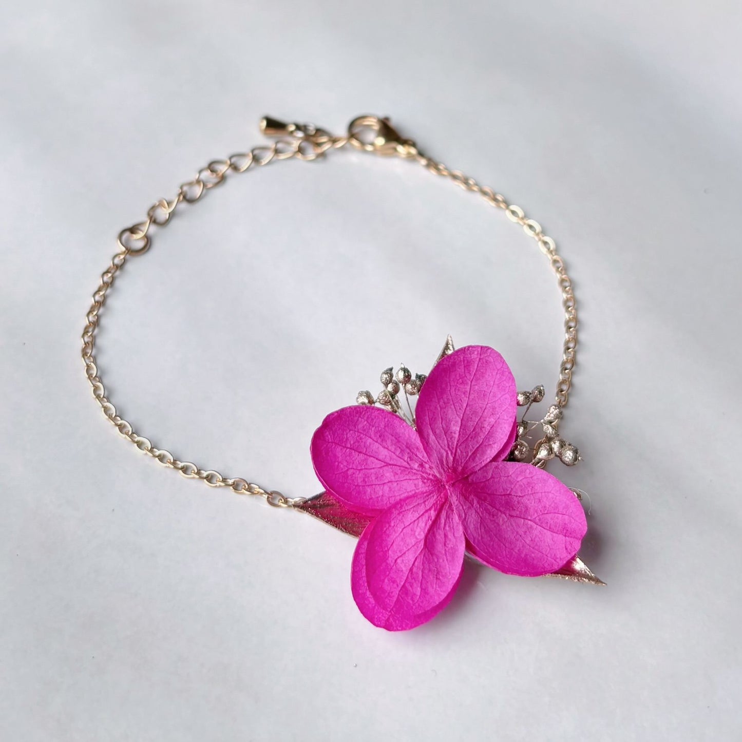 Bracelet de mariage en fleurs d’hortensias stabilisées rose fuchsia avec touches de feuillage doré, idéal pour un look bohème chic. Parfait comme cadeau pour une témoin ou une personne chère.